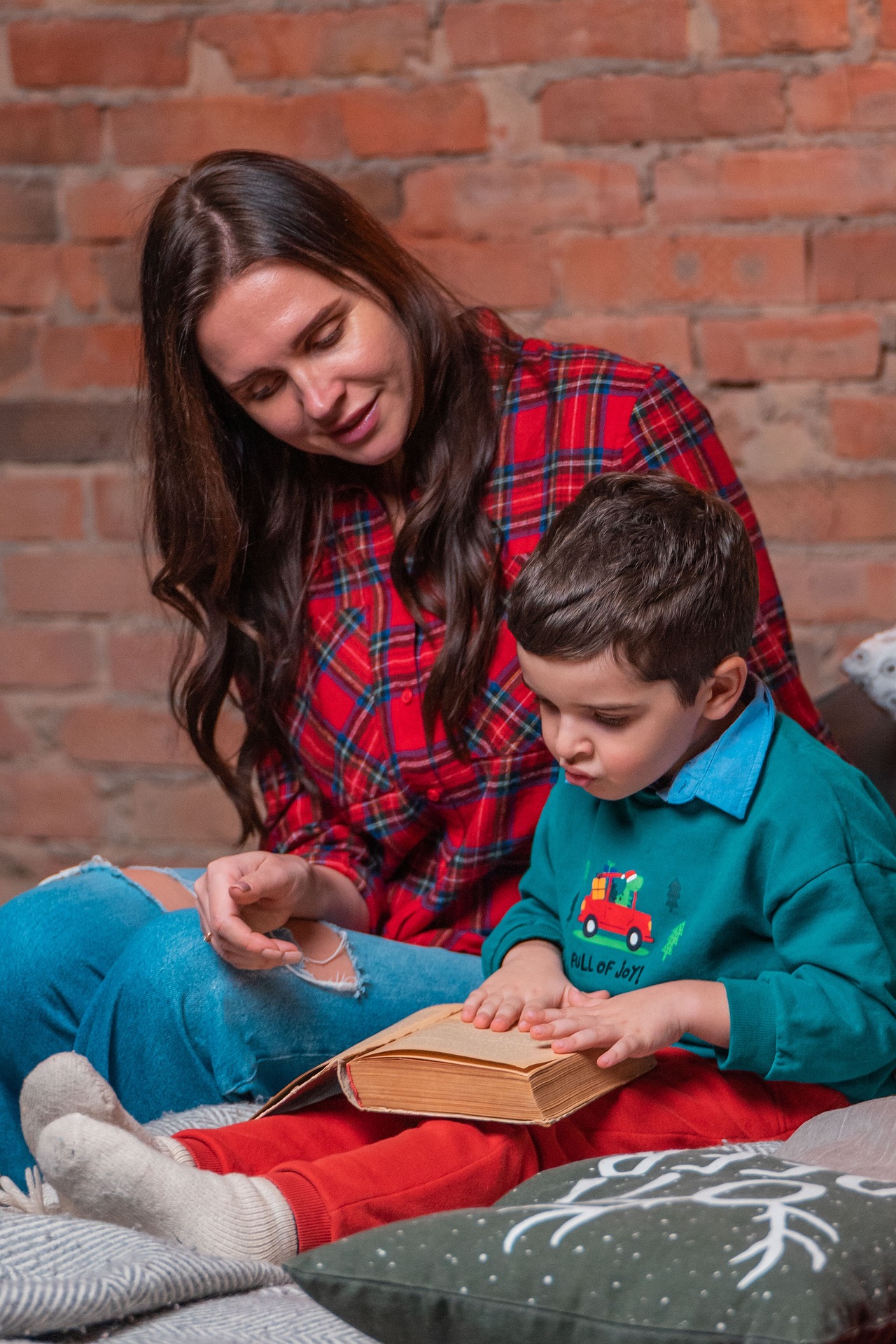 Female Adult dressed in a bright red checked shirt and green jeans reading to a young male child wearing a green long sleeved shirt
